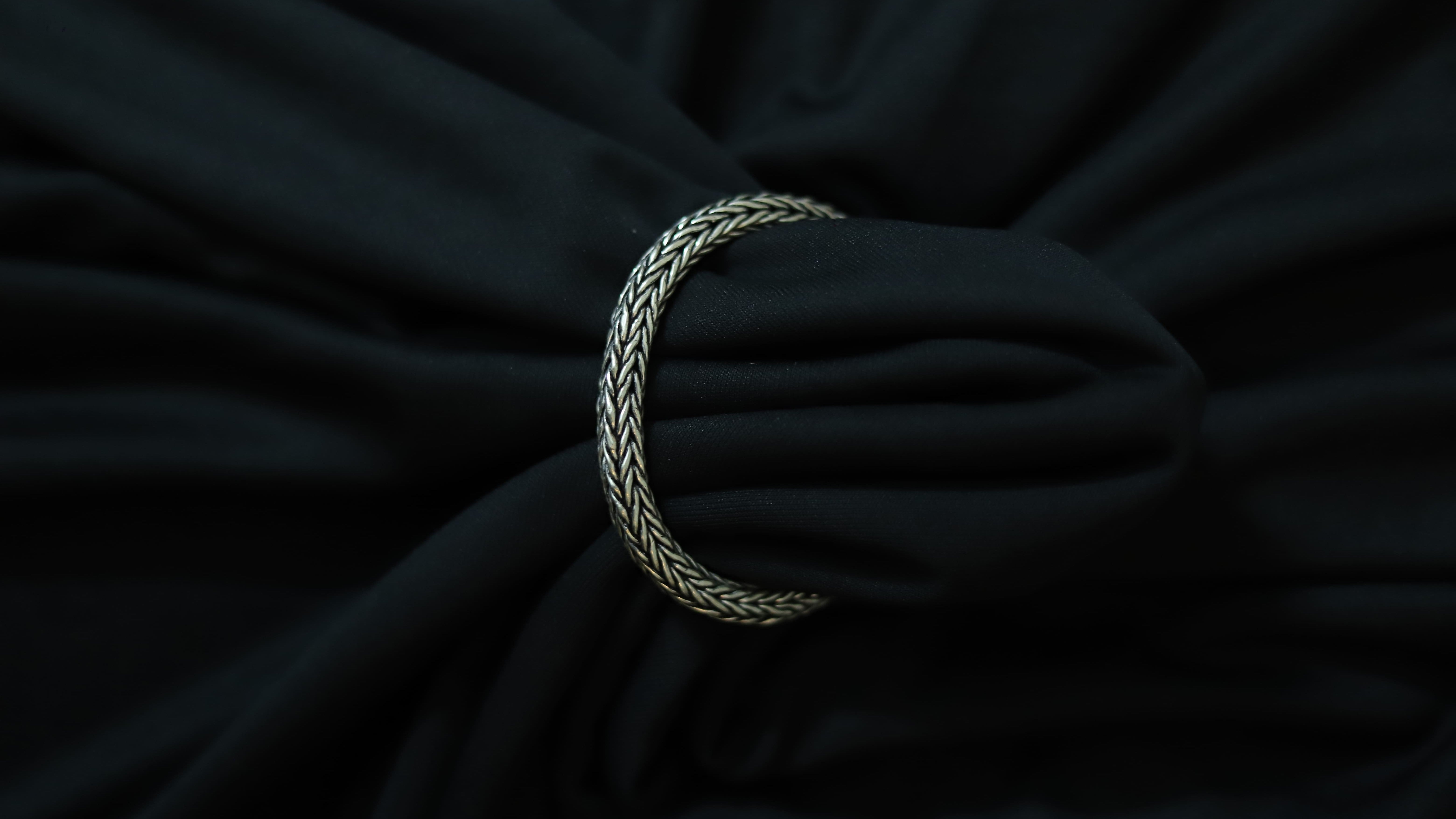 A silver braided rope bracelet or tie clip shown wrapped around a black fabric surface.